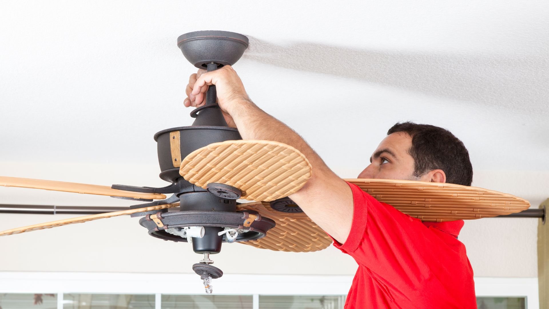 Person in red shirt installing wooden ceiling fan on white ceiling